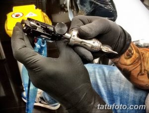 Фотография тату-машинки в руках мастера, работающего над татуировкой. Видна рука мастера в черной перчатке и детали тату-машинки.